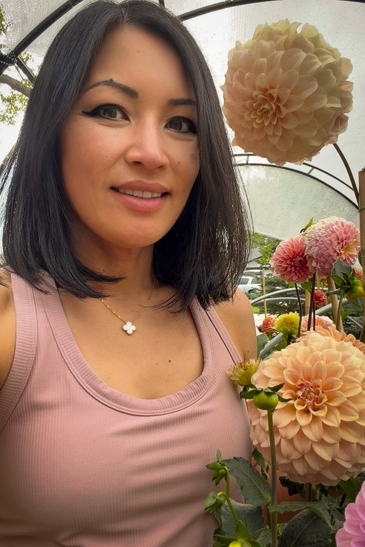Woman with shoulder-length dark hair wearing a pink tank top, standing among flowers.