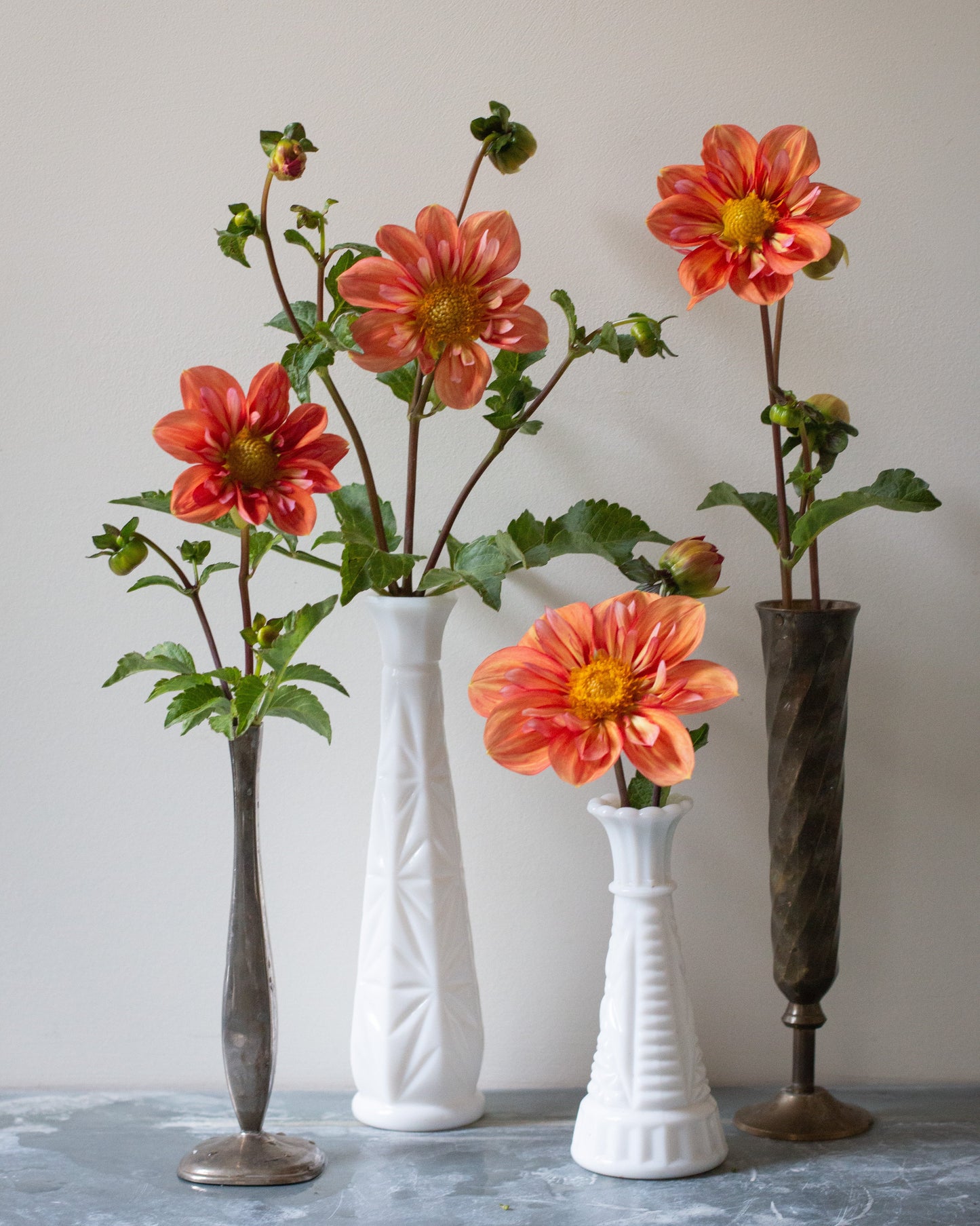 Three artificial orange dahlia flower arrangements in white and dark textured vases, set against a neutral beige wall and gray stone surface.