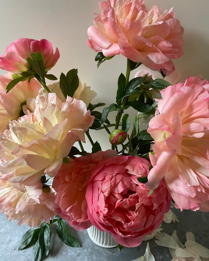 A bouquet of artificial Coral Charm peony flowers in shades of pink, peach, and cream, arranged in a small white ribbed vase on a gray surface against a light wall.