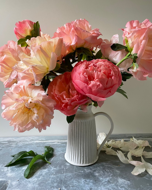 A bouquet of artificial Coral Charm peony flowers in shades of pink, coral, and pale peach, arranged in a white ribbed ceramic pitcher on a gray stone surface with scattered petals.