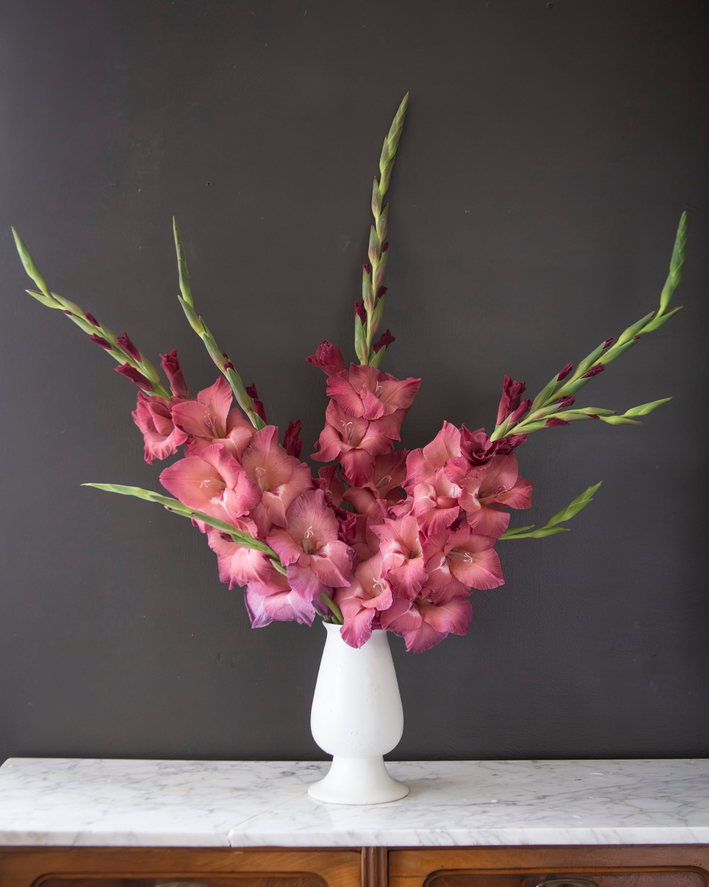 A bouquet of pink gladiolus flowers with long green stems arranged in a white pedestal vase, set on a white marble-topped wooden cabinet against a dark charcoal wall.