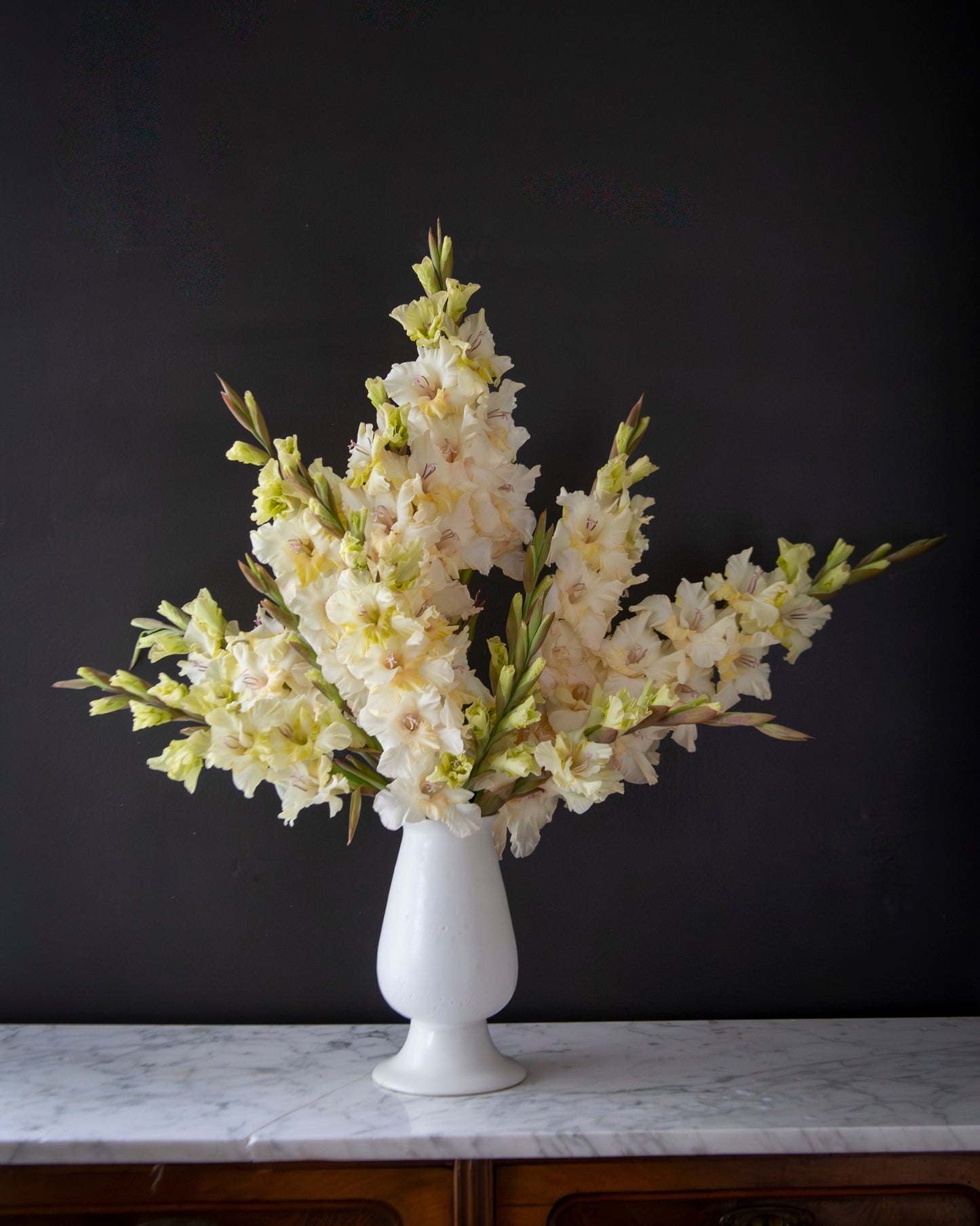 A white ceramic vase filled with a bouquet of pale cream gladiolus flowers, displayed on a white marble surface against a dark charcoal wall.