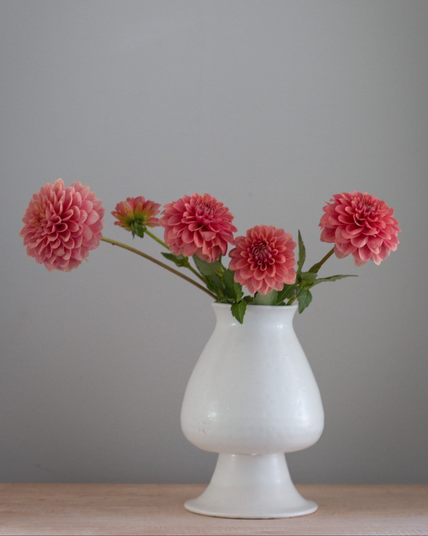 Four pink dahlia flowers with green stems arranged in a white pedestal vase on a light wood surface against a plain gray wall.