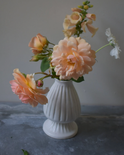 A white footed carved vase with vertical ridges, filled with pink and yellow flowers, placed on a grey surface.