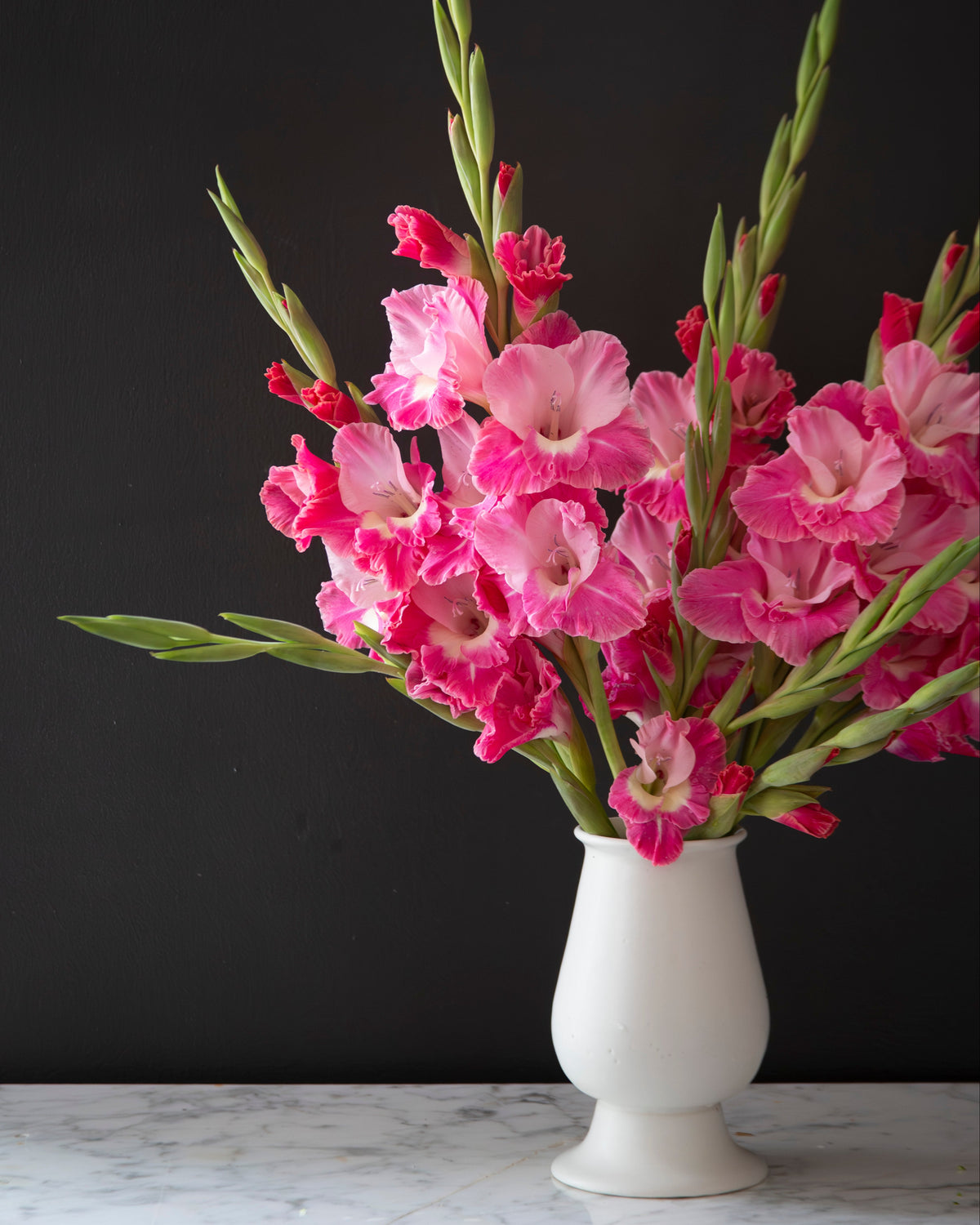 A bouquet of pink and magenta gladiolus flowers with green stems arranged in a small white ceramic vase, set against a dark charcoal background on a light marble surface.