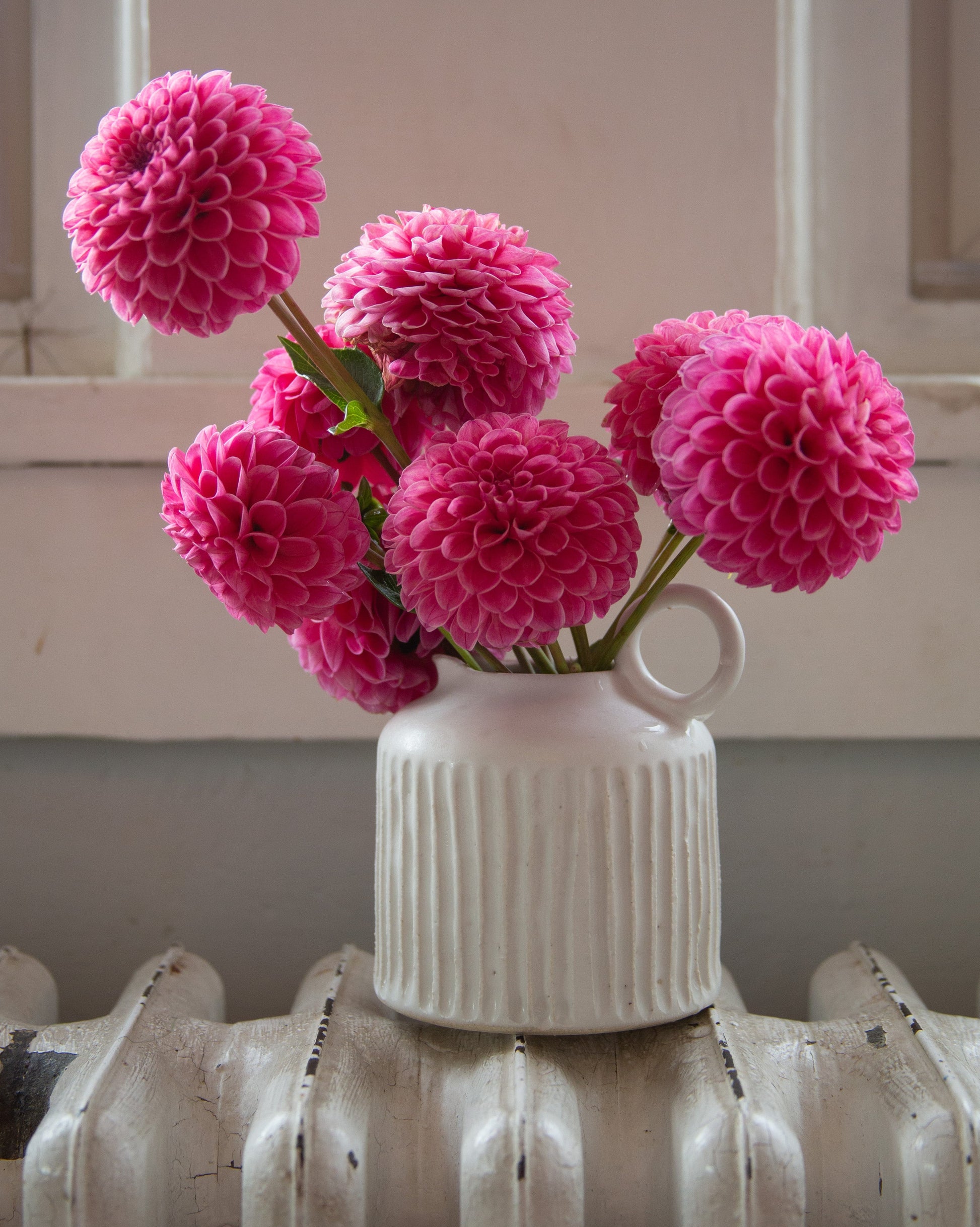 White vase with pink flowers on a radiator against a neutral wall.